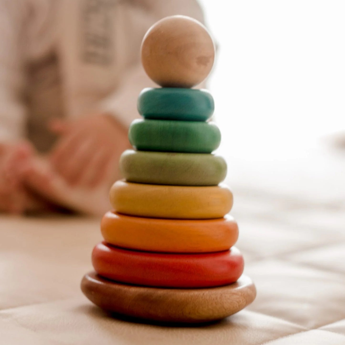 Colorful QToys wooden stacking toy on a soft surface with a blurred child in the background.